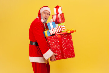 Astonished elderly man with gray beard wearing santa claus costume holding heavy stack of presents, looking at camera with open mouth, winter holidays. Indoor studio shot isolated on yellow background