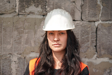 Young woman in white hard hat and orange high visibility vest, long dark hair, looking into camera. Blurred old construction site wall background