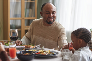 African dad helping his little daughter to drink juice from glass while they having dinner at table
