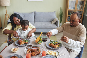 African woman feeding her child with roast chicken during dinner at table in living room
