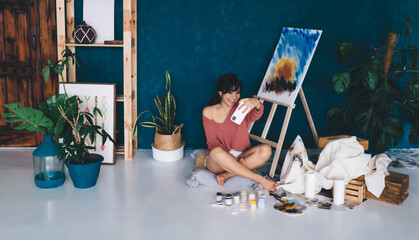 Carefree young female artist posing for smartphone camera selfie near easel and paints showing in blog her hobby. Creative smiling brunette woman photographing herself near draw oil picture