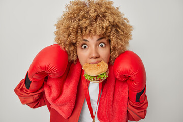 People sport and healthy lifestyle concept. Impressed shocked sportswoman eats fast food wears boxing gloves prepares for fight isolated over grey background. Female boxer with sandwich n mouth