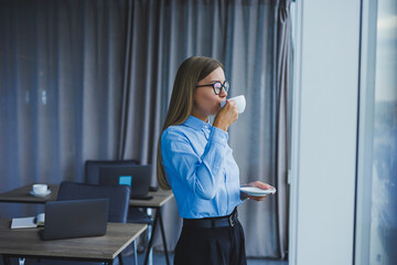 Image of a happy young woman in a classic shirt and glasses smiling and drinking coffee while standing by the window in a modern office with large windows. Remote work