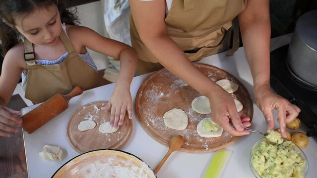 Top view mom and daughter cook together. Pretty little girl rolls out dough and sprinkles with flour while her mother fills with mashed potatoes, cooking dumplings together in summer country kitchen
