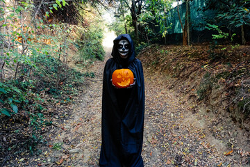 Young woman with halloween paint face mask wearing black hood - Scary witch holding spookey craved...