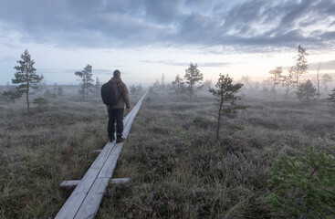 Man with the backbag and in hiking clothing stands on the nature-trail boardwalk in Nigula bog, Estonia and admiring the frosty and foggy sunrise morning view