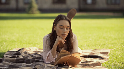 Brunette woman reading book while relaxing on blanket in park.