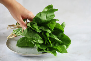 A bunch of spinach leaves lies on a plate on a marble table. Woman's hand takes spinach