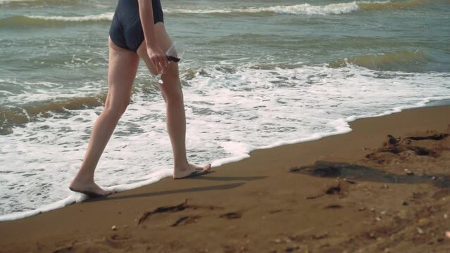 Young slender beautiful woman in elegant black bikini walks along seashore with glass of wine. Summer. Travel, weekends, leisure and lifestyle.
