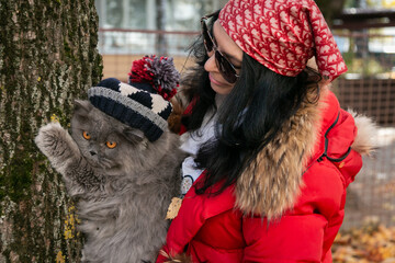 A girl in sunglasses, a headscarf and a red jacket helps a British cat in a pom-pom hat climb a tree. Love to the animals. Walk with a cat. Close-up. Blurred background.
