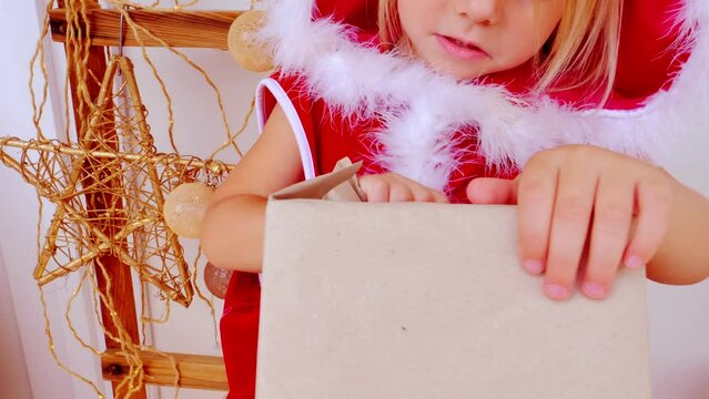 gift with purple ribbon ​​in hands of toddler close-up, small child, blonde girl 3 years old in red New Year's dress, gift from Santa Claus for Christmas, happy childhood, dream of fairy tale, magic