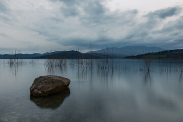 A long exposure photo of a big rock in a lake, beneath and overcast sky - 528562876