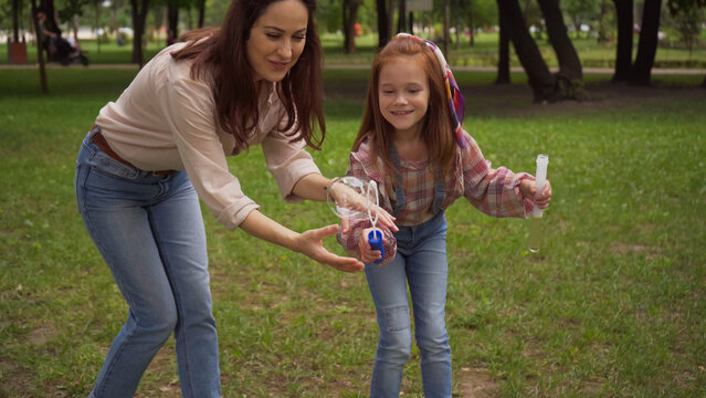 Smiling parent and girl holding soap bubbles in summer park.