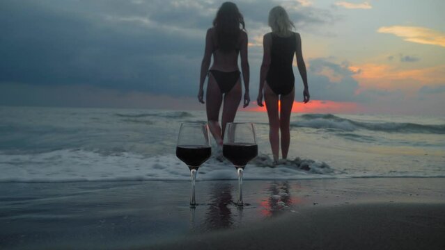 Romantic evening with stunning sunset, two women entering scream at sunset, against backdrop of two glasses of red wine and sea waves. Beautiful landscape on vacation or resort. Pair.