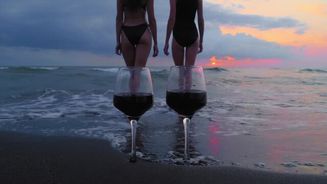 Romantic evening with stunning sunset, two women entering scream at sunset, against backdrop of two glasses of red wine and sea waves. Beautiful landscape on vacation or resort. Pair.