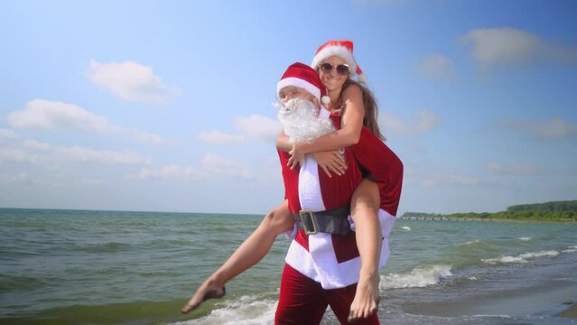 Christmas holiday. Sexy woman in Santa hat riding Santa Claus on beach by sea on hot day. They are happy. funny and happy. Hot weather, warm and exotic climate. Stay. New year.