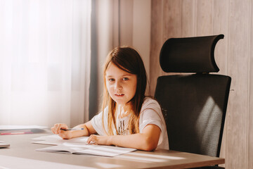 Little girl with long hairs sitting at table, holding pen, writing, learning at home. Teenager studying indoors at room with modern black chair.