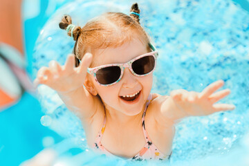Cute toddler girl wear sunglasses having fun in swimming pool with blue clean water. Family vacation in luxury resort.