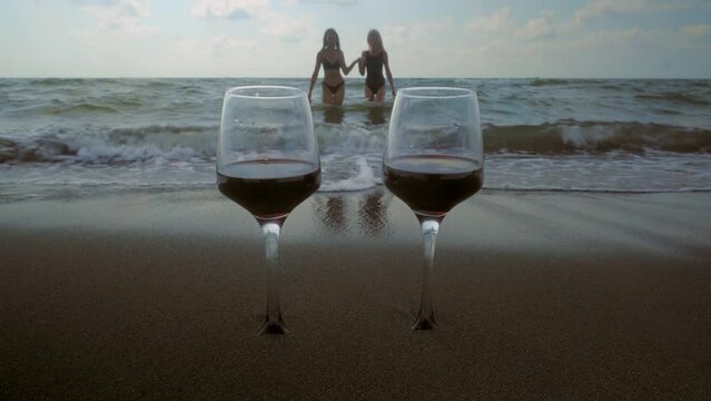 Romantic evening with stunning sunset, two women entering scream at sunset, against backdrop of two glasses of red wine and sea waves. Beautiful landscape on vacation or resort. Pair.