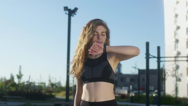 young woman with curly hair at dawn goes in for sports on street and looks at her watch. fit girl is doing workout outdoors and watching her pulse or route on smart watch.