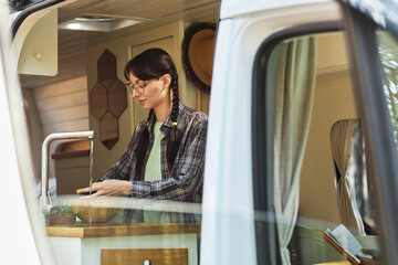 Young woman in eyeglasses washing vegetables under water in sink while standing in the van during...