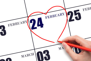 A woman's hand drawing a red heart shape on the calendar date of 24 February. Heart as a symbol of...