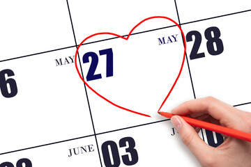 A woman's hand drawing a red heart shape on the calendar date of 27 May. Heart as a symbol of love.