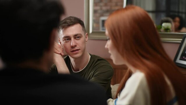Close-up side view of young redhead woman sitting around table with friends, telling story, hang out with men and women, enjoying free time. Three friends have fun at home, chatting, telling stories.