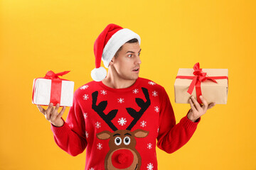 Surprised handsome man in Santa hat holding gift boxes on yellow background