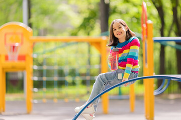 Adorable smiling little girl, 5-6 years old, sitting with a plastic bottle of water on the playground