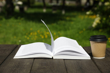 Open book and paper coffee cup on wooden table outdoors