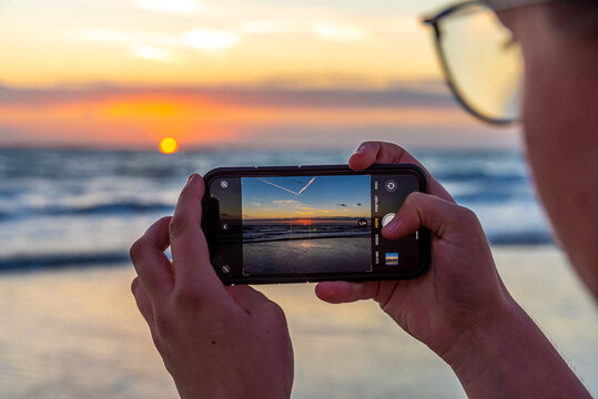 Close-up of a girls's hands with a smartphone taking photos of a bright sunset at the sea.