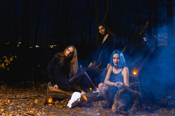 Three witches practice witchcraft in a dark forest during Halloween. A witch with blue hair hangs...