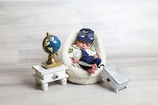 toddler boy in cute pilot costume lying at soft tiny chair little children props travel suitcase globe statue on table