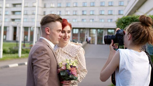 professional photographer in white T-shirt photographs bride and groom. groom grimaces at camera and makes faces. Wedding day of happy couple in park at daytime.