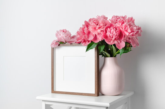 Wooden square frame mockup with pink peony flowers in white room interior, blank mockup with copy space