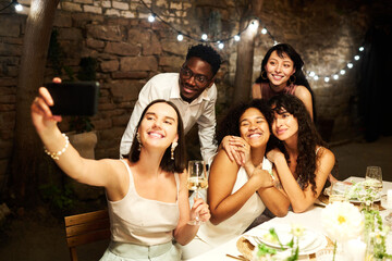 Group of happy young intercultural friends making selfie by served festive table while enjoying wedding party in luxurious restaurant