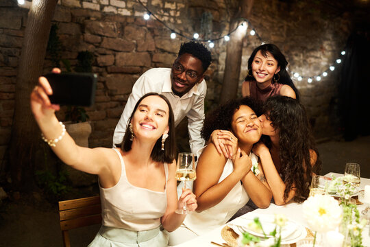 Young brunette woman taking selfie with group of friends and affectionate girl kissing her bride on cheek at wedding party