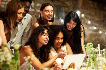 Two young intercultural brides and their friends communicating in video chat while sitting by served festive table during wedding feast