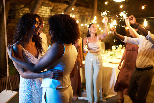 Two young amorous intercultural women in white wedding attire dancing in front of camera against their friends with sparklers having fun