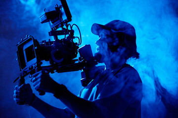 Bearded videographer in baseball cap holding steadicam while standing in smoke and making all the...