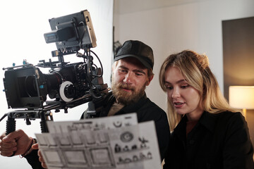 Bearded videographer with videocamers and his blond female assistant discussing working documents...