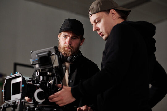 Two serious men in black sweatshirta discussing settings of steadicam during commercial shooting while one of them consulting another
