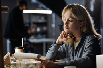 Young serious blond woman concentrating on reading information in document while sitting by workplace in production studio