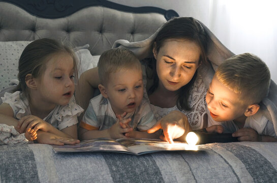 Mom with a flashlight reads a book to children covering them with a blanket.