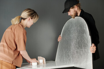 Young blond woman in beige t-shirt bending over table while making composition in front of...