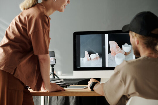 Close-up of young photographer and his assistant by workplace with computer monitor during discussion of new photos