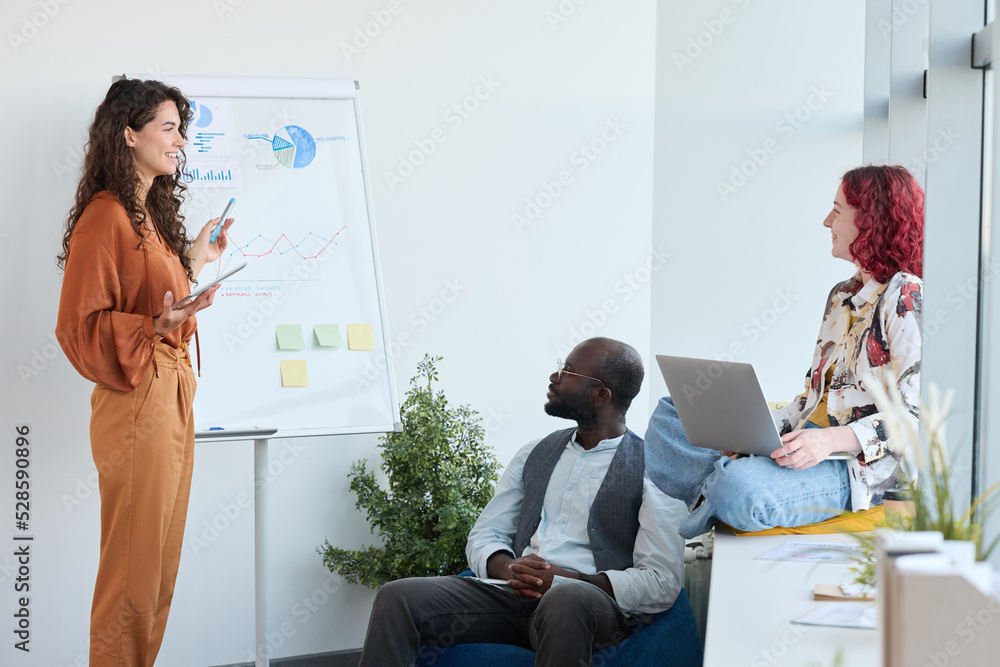 Poster young confident businesswoman standing by whiteboard with financial diagrams and charts while explai