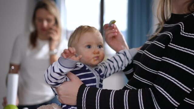 Side view unrecognizable Caucasian woman feeding baby girl with blurred lady talking on phone at background. Young mother taking care of child at home meeting with friend. Multitasking and motherhood