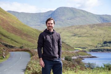 Caucasian man standing with hand in pockets on the side of Ben Gorm mountain, on Road R335, County Mayo, Ireland. Lake, road and mountains on background